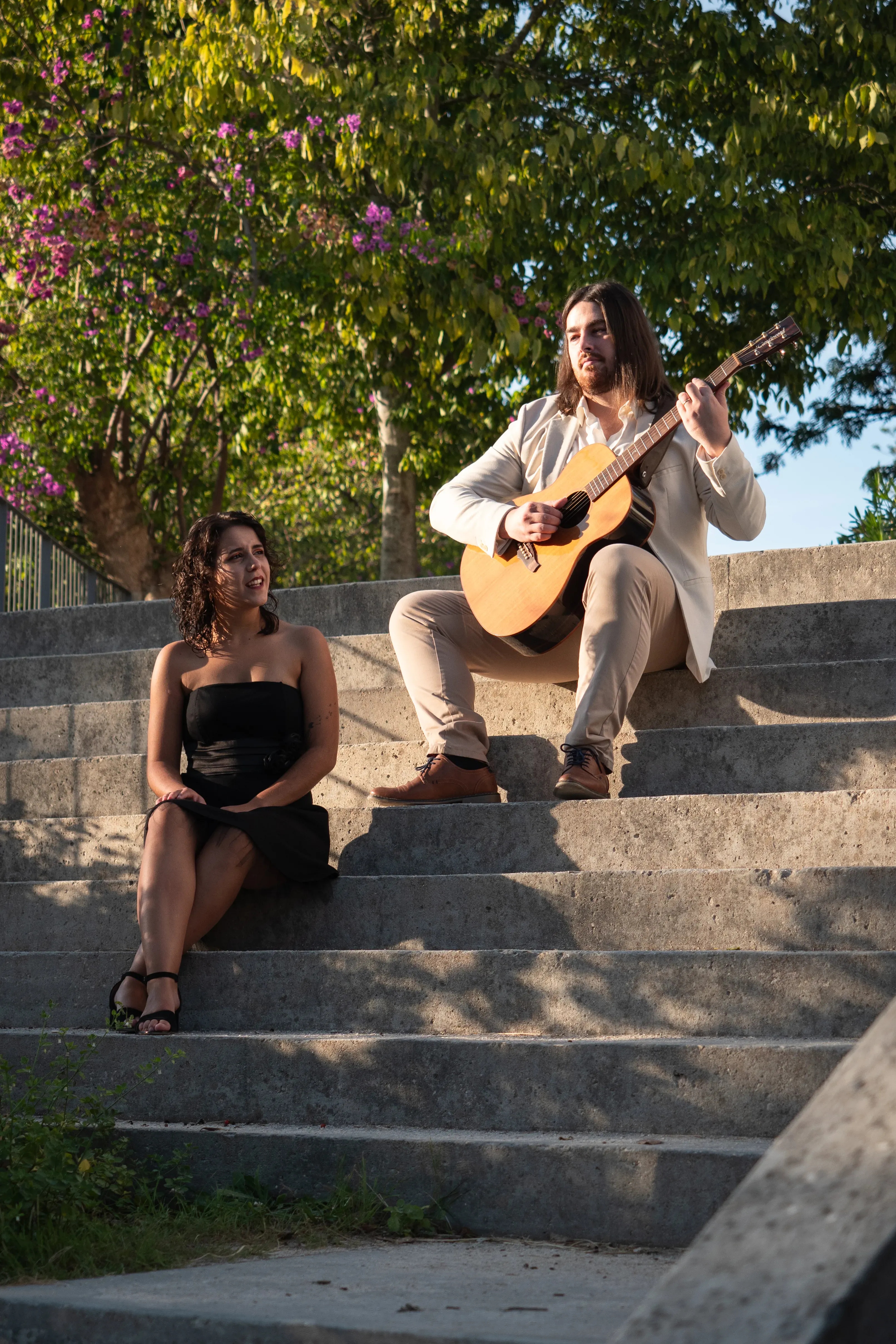 Numa escadas de betão de um parque, um casal está sentado. O rapaz está em cima a tocar guitarra clássica, tem cabelo comprido liso e está vestido com um fato claro. A rapariga, mais em baixo, está a cantar e vestida com um vestido preto e tem cabelo encaracolado curto.